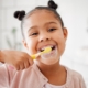 little girl with two tiny buns on her hair smiling as she brushes her teeth with a yellow toothbrush