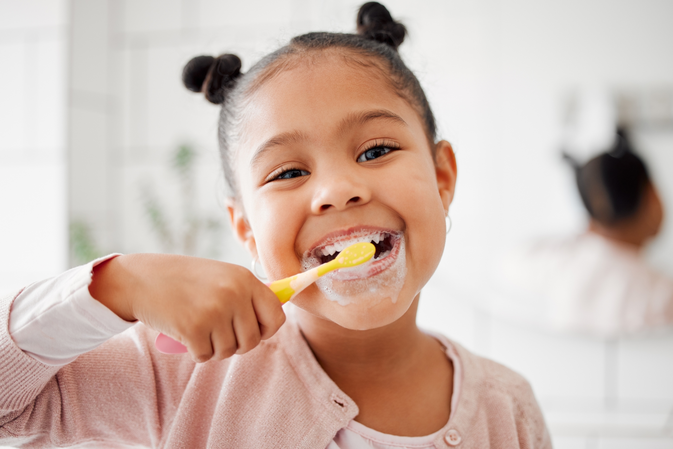 little girl with two tiny buns on her hair smiling as she brushes her teeth with a yellow toothbrush